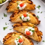 A serving plate featuring baked chicken tacos filled with shredded chicken, topped with dollops of sour cream and small pieces of diced tomato.