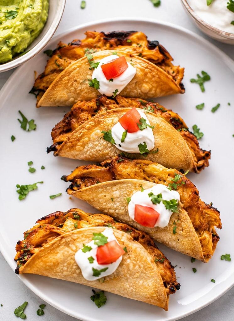 A serving plate featuring baked chicken tacos filled with shredded chicken, topped with dollops of sour cream and small pieces of diced tomato.