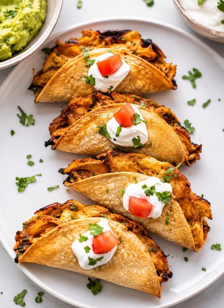 A serving plate featuring baked chicken tacos filled with shredded chicken, topped with dollops of sour cream and small pieces of diced tomato.