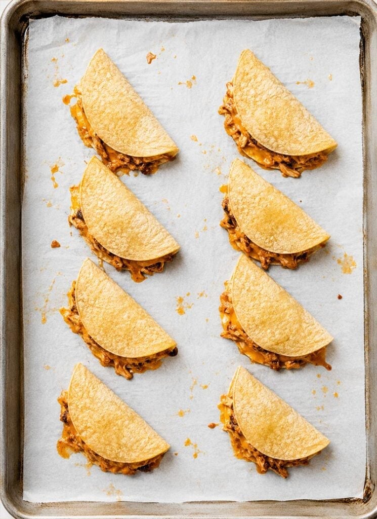 A baking sheet lined with parchment paper holds eight partially assembled taco shells filled with shredded chicken and cheese.